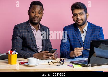 Shot of two businessmen wearing suits sitting in office at desk, using laptop and cellphone. Indian man sharing ideas with african american colleague, Stock Photo