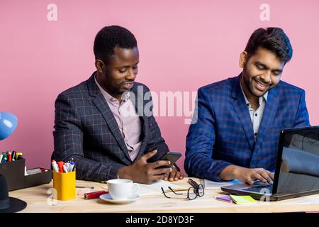 two businessmen wearing suits sitting in office at desk, using laptop and cellphone. Indian man sharing ideas with african american colleague, showing Stock Photo