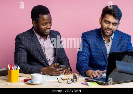 Shot of two businessmen wearing suits sitting in office at desk, using laptop and cellphone. Indian man sharing ideas with african american colleague, Stock Photo