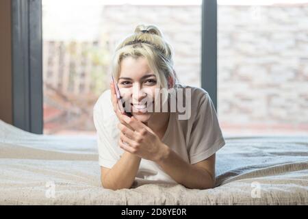 Close-up portrait of a nice sweet winsome attractive charming cute lovely adorable stunning cheerful blond lady lying on bed taking b y phone Stock Photo
