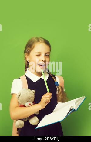 Study and back to school concept. Girl with braids winks with happy face. Kid in school uniform isolated on green background. Pupil holds blue book, marker and teddy bear Stock Photo