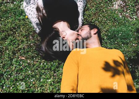 Beautiful young couple lie on the green grass in a spring meadow in the park top view. A beautiful woman lies and kisses a man on the ground. Relation Stock Photo