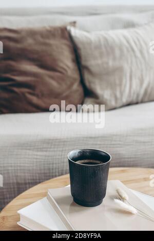 Books, dry bunny tail grass and black cup of coffee on round wooden table. Blurred beige sofa background with linen cushions. Hygge, cozy lifestyle Stock Photo