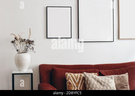 Portrait wooden and black picture frames mockup on white wall. Modern white ceramic vase with dry grass on marble table. Rusty velvet sofa with linen Stock Photo