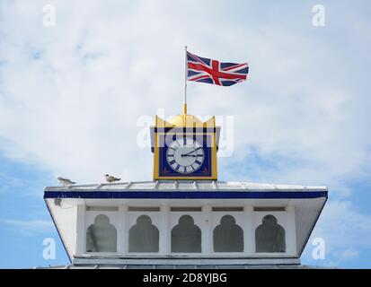 The Union Jack flag flies atop the clock tower of Eastbourne pier. Gulls sit on the roof in sunshine. Stock Photo