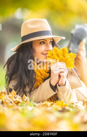 Gorgeous brunette holds yellow fallen maple leaves looking to the side while lying on the ground outdoors. Stock Photo