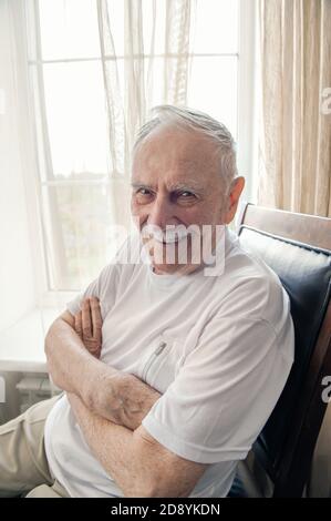 old man is sitting in a chair. Smiling old man sitting in a chair by the window, portrait, close-up Stock Photo
