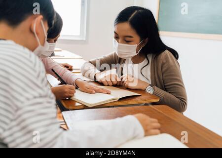 Close-up of Asian female teacher wearing a face mask in school building tutoring a primary student children. Elementary pupils writing and learning in classroom. Covid-19 school reopen concept Stock Photo
