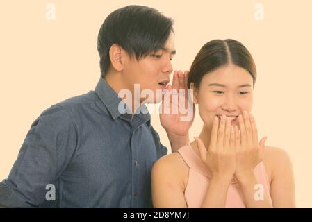 Studio shot of young Asian couple with man whispering to woman laughing Stock Photo