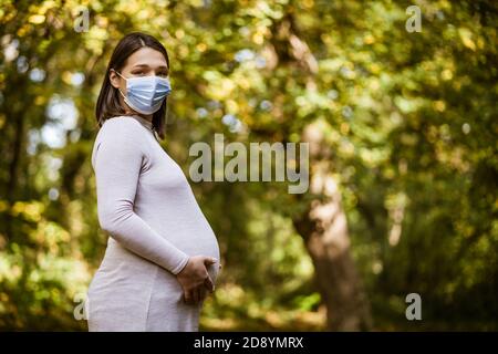 Portrait of pregnant woman in park in autumn. She is wearing protective face mask. Covid-19 pandemic concept. Stock Photo