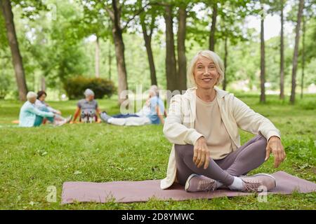 Full body shot of happy senior woman with gray hair sitting on mat in park with legs crossed smiling at camera Stock Photo