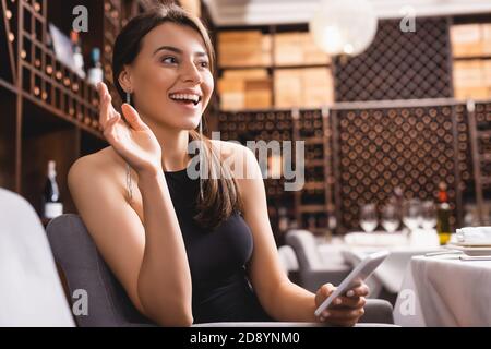 Selective focus of elegant woman waving hand while holding smartphone in restaurant Stock Photo