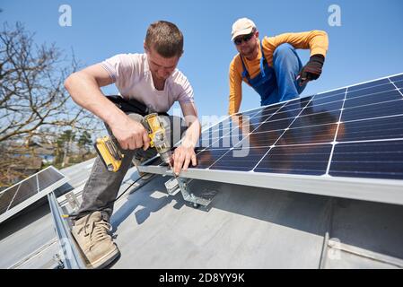 Male engineers installing solar photovoltaic panel system using screwdriver. Two electricians mounting solar module on roof of modern house. Alternative renewable innovation concept. Stock Photo