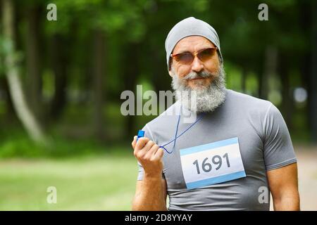 Waist up portrait shot of modern bearded mature adult man wearing sports outfit holding whistle looking at camera Stock Photo