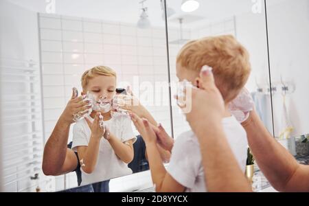 Father with his son is in the bathroom have fun by using shaving gel and looking in the mirror
