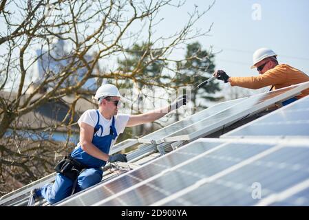 Male engineers installing solar photovoltaic panel system. Electricians mounting blue solar module on roof of modern house. Alternative energy ecological concept. Stock Photo