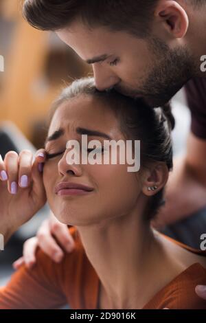 responsive man kissing head of crying girlfriend wiping tears with hand Stock Photo
