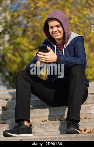 Smiling teenager watching funny video on cellphone screen or chatting with friends online. Attractive caucasian teenage boy in hood sitting on stairs Stock Photo
