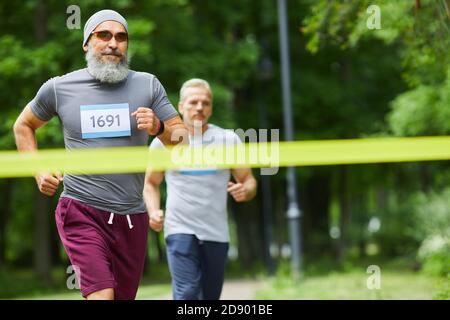 Two active sporty senior men finishing running marathon race with bearded man being first, medium long shot Stock Photo