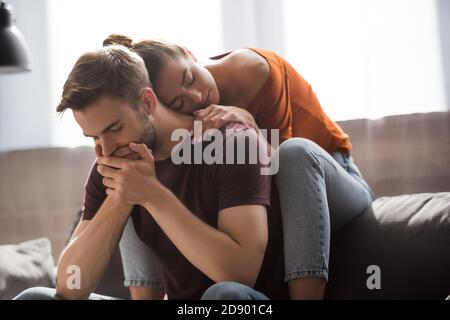 young woman leaning on offended boyfriend sitting on sofa with bowed head and covering mouth with hands Stock Photo