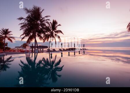Beautiful poolside and sunset sky. Luxurious tropical beach landscape, deck chairs and loungers and water reflection. Amazing sunset infinity swimming Stock Photo