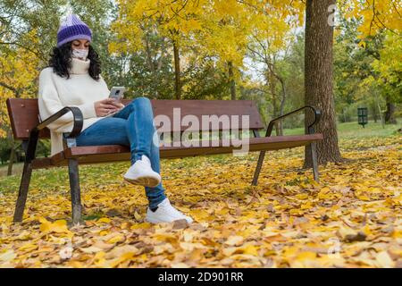young woman with a protective face mask due to the global coronavirus pandemic, covid-19 is sitting on a bench in a park in autumn chatting with her m Stock Photo