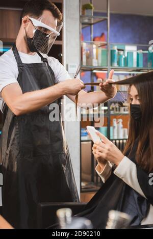 hairdresser in protective equipment cutting hair of woman using smartphone, blurred background Stock Photo