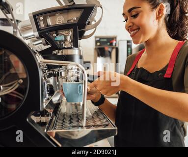 Smiling female barista using coffee maker in cafeteria. Woman in apron preparing espresso. Stock Photo