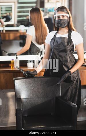 hairdresser in apron and face shield holding scissors and comb while pointing with finger at armchair on blurred background Stock Photo