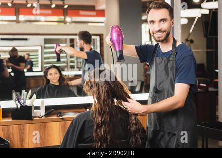 cheerful hairdresser drying hair of client while looking at camera near blurred mirror reflection Stock Photo