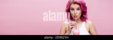 surprised young woman with colorful hair drinking strawberry milkshake from straw isolated on pink, panoramic shot Stock Photo