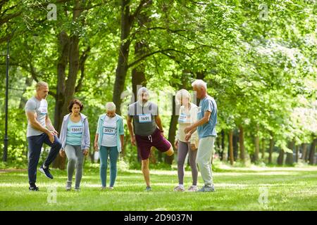 Long shot of active seniors taking part in summer marathon race standing together somewhere in park doing stretching exercise Stock Photo