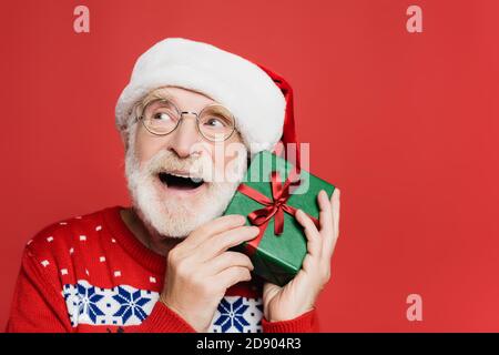 Dreamy senior man in santa hat holding present isolated on red Stock Photo