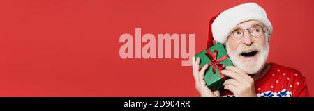 Dreamy senior man in eyeglasses and santa hat holding gift box isolated on red, banner Stock Photo