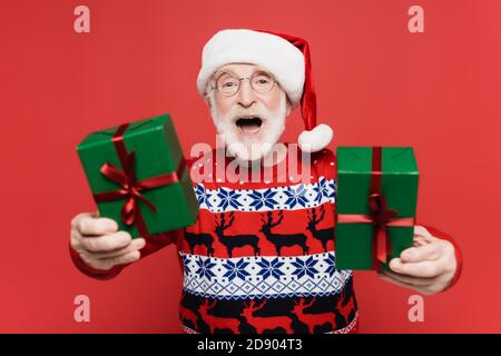 Cheerful elderly man in santa hat holding gift boxes on blurred foreground isolated on red Stock Photo