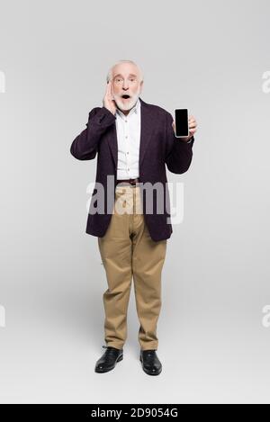 Shocked elderly man with hand near head holding smartphone with blank screen on grey background Stock Photo
