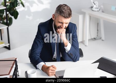 Businessman yawning while working near laptop and papers on table Stock Photo