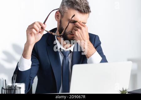Tired businessman holding eyeglasses while working near laptop on blurred foreground Stock Photo
