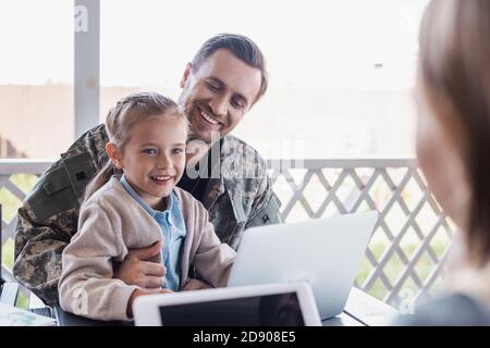 Happy girl sitting with military serviceman near laptop with blurred woman on foreground Stock Photo