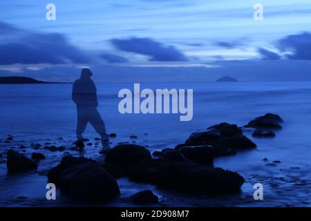 Ailsa Craig from Croy Shore, Ayrshire, Scotland, a hooded ghostly fiqure on rocks looking towards Ailsa Craig. Semi see through, a ghost from the past Stock Photo