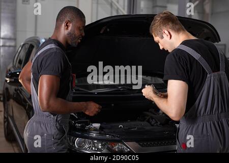 multi-ethnic team of african and caucasian men working in auto service together, friendly men in uniform repair the car hood Stock Photo