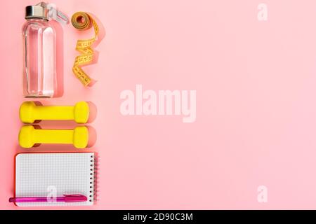 Bottle of water, dumbbells, measuring tape notebook with pen on pink background. Healthy lifestyle, Gym and workout flat lay concept. Stock Photo