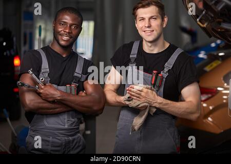 multiethnic team of two auto mechanic males at work place, successful work of diverse workers in uniform, they are posing at camera Stock Photo