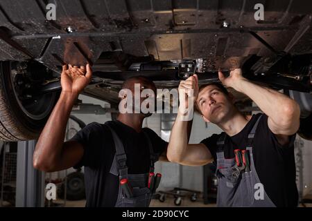 two interracial car mechanics in uniform checking car in automobile service with lifted vehicle, african and caucasian men repair a car together Stock Photo