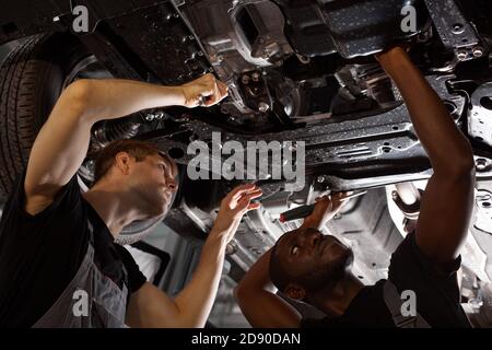 view from below on diverse men repairing car bottom, multi-ethnic cooperation, two young african and caucasian guys work together. Stock Photo