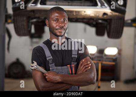 professional black auto mechanic looking at camera, handsome black guy in uniform is keen on repairing car. automobile in the background Stock Photo
