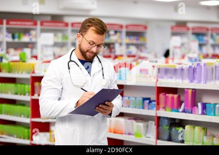 pleasant male pharmacist writing on clipboard at pharmacy, make notes, portrait of handsome bearded worker of drugstore in white gown Stock Photo