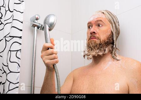 Confused bearded man with soapy head standing in the bathroom and looking at the shower while the water supply has stopped. No water coming from tap Stock Photo