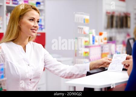 young beautiful caucasian pharmacist is working with customer in modern drugstore, serve the client, wearing white medical gown Stock Photo