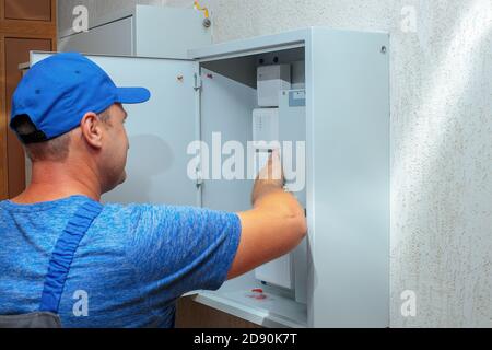 An electrician in overalls and a baseball cap examines a Cabinet with sensors and equipment.  Stock Photo